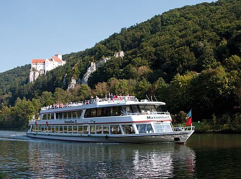 Ein voll besetztes Personenschiff fährt auf dem Main-Donau-Kanal. Über ihm ragt die Burg Prunn aus dem grünen Hängen heraus. Der Himmel strahlt wolkenlos.