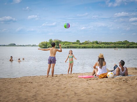 Am sandigen Badestrand sitzt eine Frau im weißen Kleid auf ihrem Badetuch, daneben liegt ein Mann. Die zwei Kinder, ein Junge mit dem Rücken zu uns und das Mädchen spielen mit einem Ball. Drei Personen plantschen im Altmühlsee.