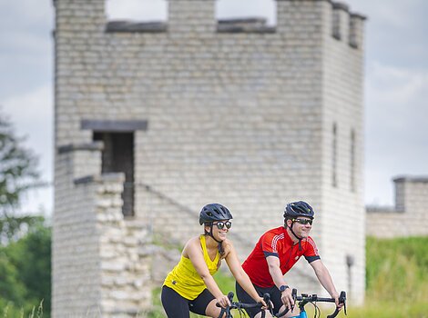 Zwei Gravelbiker, eine Frau und ein Mann, radeln am Kastell vorbei. Es ist ein Turm mit Tür zu sehen.