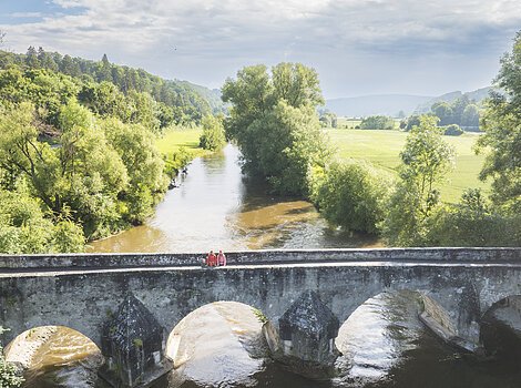 Eine alte, massige steinerne Brücke überspannt den Fluss und der Blick geht weit ins grüne Altmühltal.