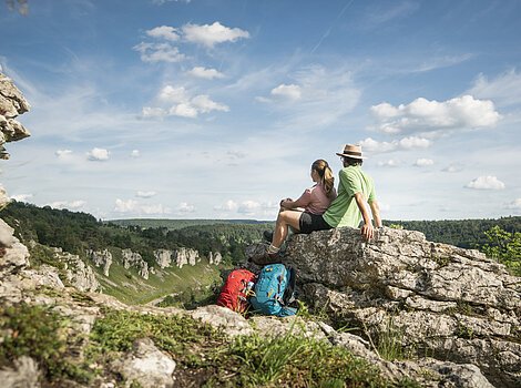 Links ist im Vordergrund Gesteinsformation im Bild. Mittig sitzt ein Mann mit Strohhut auf einem Felsen, das die zwölf Aposteln formt. Vor ihm sitzt eine Frau mit rosa T-Shirt und lehnt sich an den Mann an.  Beide blicken ins Tal und zum blauen Himmel. Vor ihnen lehnen deren Wanderrucksäcke an dem Felsen.