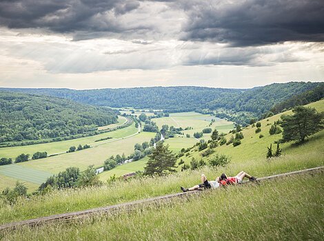 Zwei Wanderinnen liegen auf einer langen Holzbank und schauen ins weitläufige grüne Altmühltal.