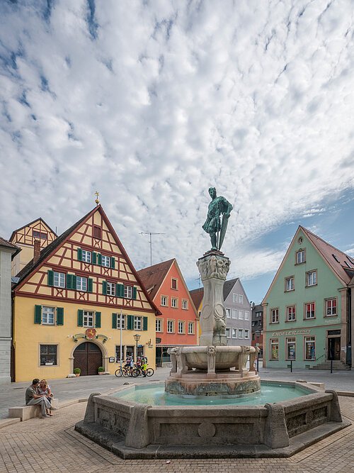 Der Kaiser-Ludwig-Brunnen am Marktplatz in Weißenburg. Auf einer Bank sitzt ein Pärchen und zwei Radler fahren gerade über den Platz. Die farbenprächtigen Häuser säumen den Platz ein.