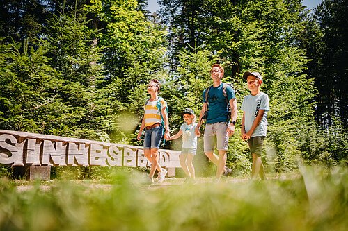 Eine Familie wandert an einem sonnigen auf einem Waldweg vor dem langgezogenem Holzschild "Sinnespfad" im Walderlebniszentrum Schernfeld.