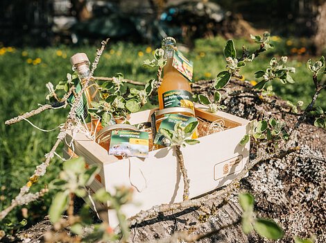 Holzkiste mit Bio-Produkten, darunter Apfelsaft, Honig und Likör, auf einem Baumstamm im Freien platziert.