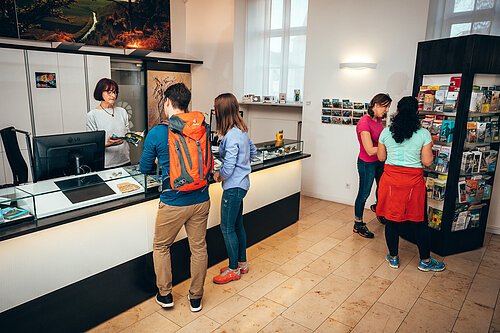 An der Tourist-Information steht ein Pärchen und lässt sich von einer Mitarbeiterin mit halblangen Haaren beraten. Zwei Frauen stehen rechts interessiert am gefülltem Prospektständer.