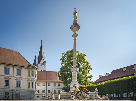 Zwei Radfahrer fahren an einer Mariensäule auf einem Platz mit historischen Gebäuden vorbei.