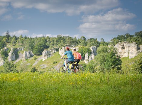 Zwei Personen fahren mit Fahrrädern auf einem grasbewachsenen Feld vor einer Felslandschaft.
