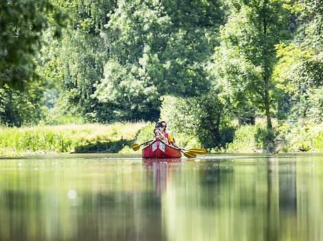 Zwei Personen paddeln in einem roten Kanu auf einem Fluss, umgeben von grünen Bäumen.