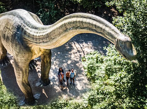 Große Dinosaurierstatue im Wald, Menschen stehen darunter und schauen nach oben.