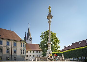 Zwei Radfahrer fahren an einer Mariensäule auf einem Platz mit historischen Gebäuden vorbei.