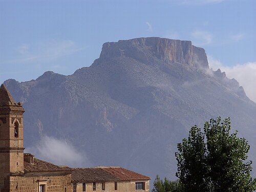 Muela - Partnernaturpark Sierra María - Los Vélez Ein Berg mit flachem Gipfel im Hintergrund, im Vordergrund ein Gebäude mit Turm und ein Baum.