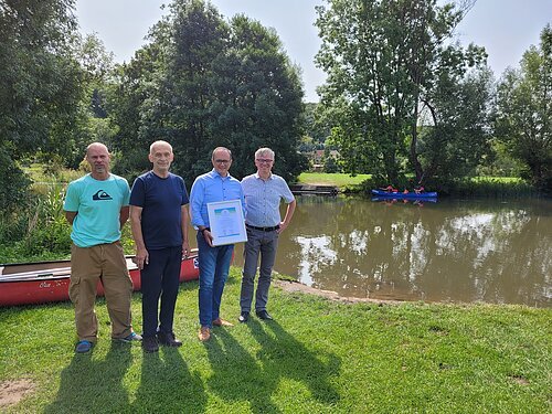 Vier Männer stehen auf einer Wiese vor einem Teich, einer hält eine gerahmte Urkunde. Im Hintergrund sind Bäume und ein Kanu auf dem Wasser.