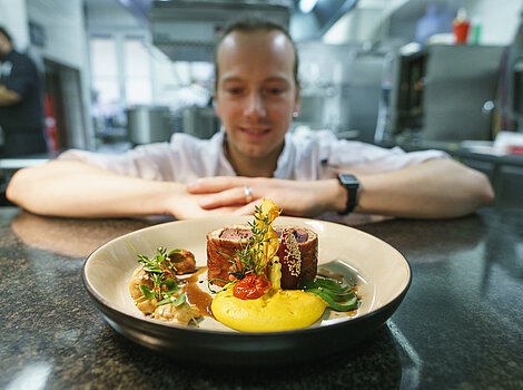 Ein Teller mit kunstvoll angerichtetem Essen auf einer Küchentheke, im Hintergrund ein unscharfer Koch.