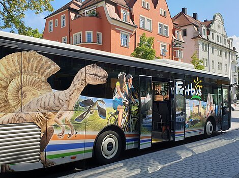 Bus mit Werbung für Freizeitaktivitäten, darunter Dinosaurier, Radfahrer und Landschaften, an einer Straße mit Gebäuden