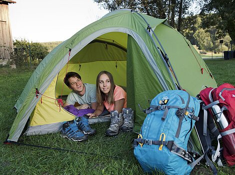 Zwei Personen liegen in einem grünen Zelt, davor Wanderschuhe und zwei große Rucksäcke auf Gras.