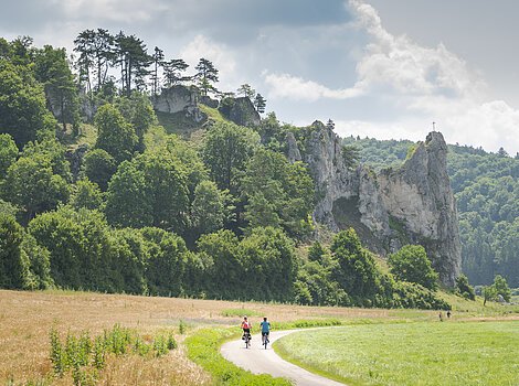 Zwei Radfahrer auf einem Weg vor bewaldeten Felsen mit einem Kreuz auf dem höchsten Felsen.