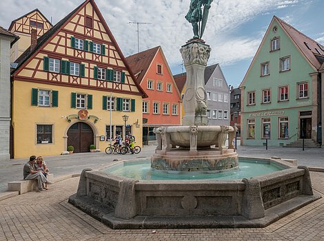 Brunnen mit Statue auf einem Platz, umgeben von Fachwerkhäusern und Menschen, unter bewölktem Himmel.