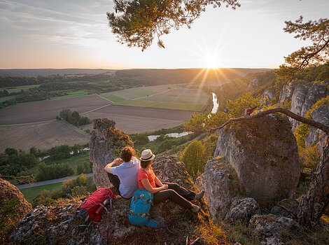 Zwei Wanderer sitzen auf Felsen mit Blick auf ein Tal und die untergehende Sonne am Horizont.