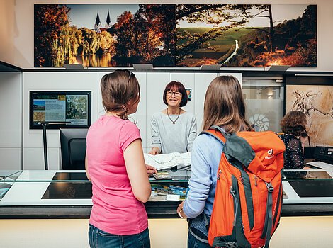 Informationszentrum Naturpark Altmühltal Zwei Frauen mit Rucksack und pinkem Shirt erhalten an einem Informationsschalter eine Karte von einer Mitarbeiterin.