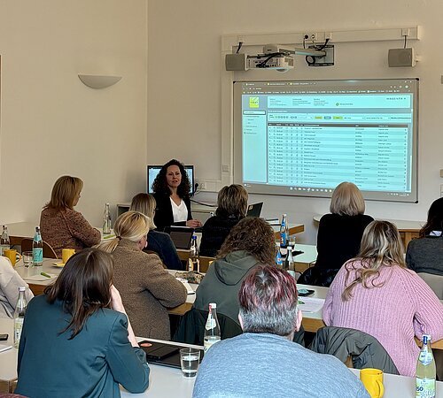 Frau hält Schulung vor Publikum, Präsentation mit Tabellen auf Leinwand in hellem Raum mit Tischen und Getränken.