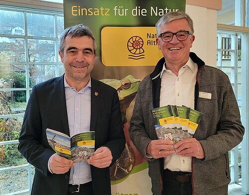 Neue Broschüre „Die schönsten Ausflugsziele“ Zwei Männer stehen vor einem Banner mit der Aufschrift „Einsatz für die Natur“ und halten Broschüren vom Naturpark Altmühltal.