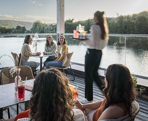 Terrasse des Restaurants ‚Mein Seeblick‘ am Kratzmühlsee Fünf Frauen sitzen auf einer Terrasse am Fluss, Kellnerin bringt Getränke auf Tablett, Wald im Hintergrund.