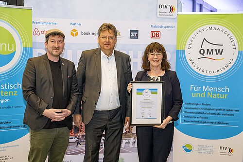Angelika Schäffer nimmt Auszeichnung entgegen. Drei Personen stehen vor Bannern des Qualitätsmanagements Wassertourismus, eine Frau hält eine Urkunde.