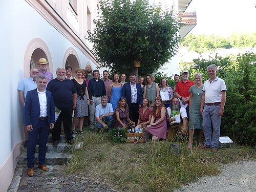 Gruppenfoto von 26 Personen vor einem weißen Gebäude mit Bäumen und Gras im Vordergrund, einige halten Zertifikate und eine Holzkiste.