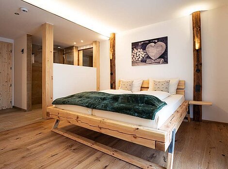 Schlafzimmer mit Holzbett, grünem Überwurf, Holzboden und offener Dusche hinter Holztrennwand.