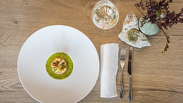 Weißer Teller mit kleiner, kunstvoll angerichteter Speise, Besteck, Glas mit Getränk und Dekoration auf Holzoberfläche