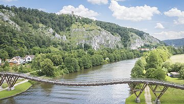 Fluss mit hölzerner Fußgängerbrücke, bewaldeten Hügeln und Häusern im Hintergrund bei bewölktem Himmel.