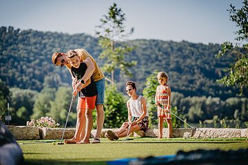 Vater zeigt Sohn das Golfspielen, Mutter sitzt im Hintergrund, Tochter steht mit Golfstock auf Rasenfläche.