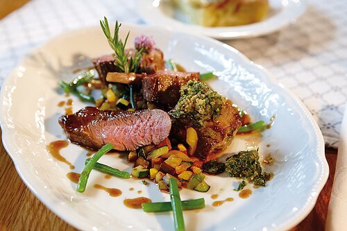 Auf einem weißen Teller liegt rosa gebratenes Lammfleich mit grünen Bohnen und Ratatouillegemüse, garniert mit Kräutern.