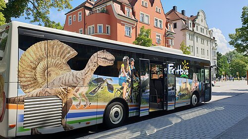 Bus mit Werbung für Freizeitaktivitäten, darunter Dinosaurier, Radfahrer und Landschaften, an einer Straße mit Gebäuden