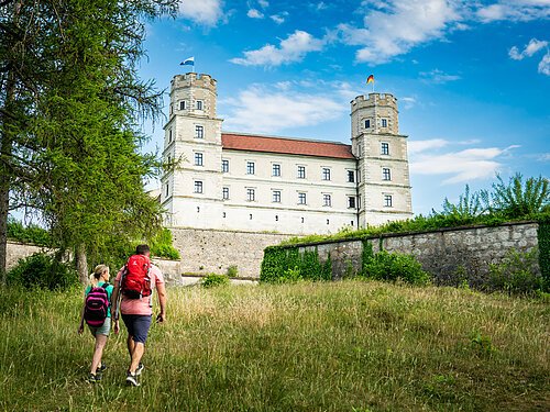 Eine Frau und ein Mann laufen leicht bergauf auf die Fassade einer Burg mit zwei Türmen zu. Sie tragen kurze Hosen, T-Shirts und je einen Rucksack.