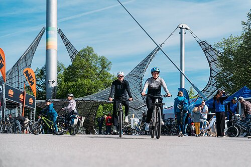 Zwei Personen fahren mit E-Bike auf dem Gelände des Olympiapark Fahrrad. Um sie herum sieht man Messebesucher der E BIKE DAYS München.