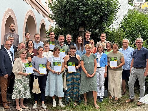 23 Naturparkführer erhalten Zertifikat Gruppenfoto von 26 Personen, einige halten Zertifikate, im Freien vor einem Gebäude mit Bögen und Bäumen.
