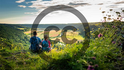 Zwei Wanderer mit Rucksäcken sitzen auf einem Hügel und blicken auf ein Tal mit Feldern und Wald.Über dem Bild befindet sich als Wasserzeichen das Copyright-Symbol.