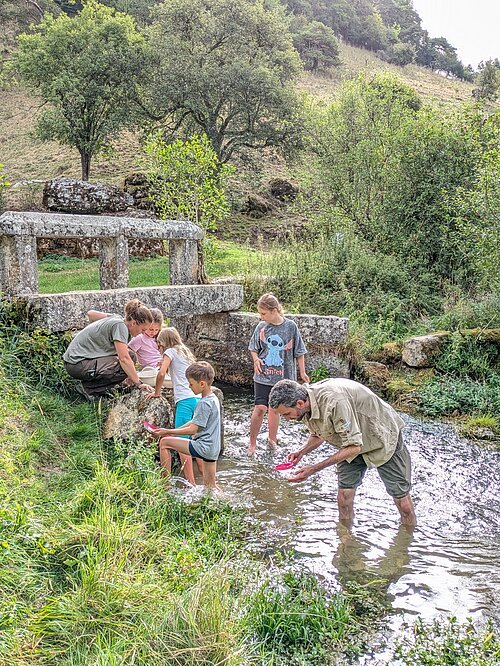 Sechs Personen, darunter Kinder und Erwachsene, stehen in einem flachen Bach vor einer Steinbrücke in einer grünen Landschaft.