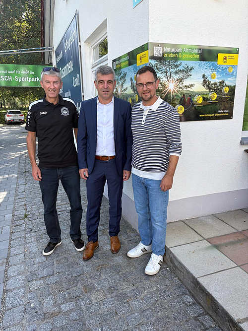Drei Männer stehen nebeneinander vor einer weißen Hauswand mit einem Naturpark Altmühltal Infoschild im Hintergrund.