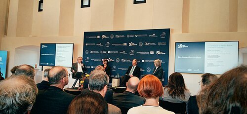 Podiumsdiskussion mit vier Personen auf Bühne vor Publikum in einem Saal mit zwei Bildschirmen und Banner im Hintergrund.